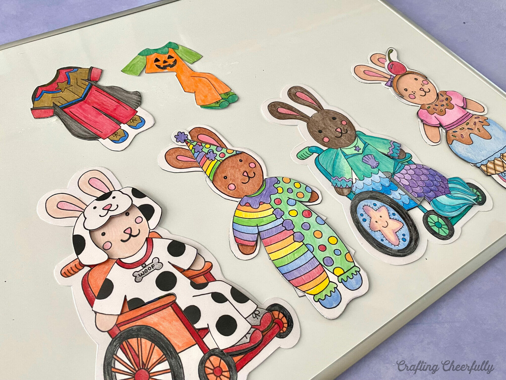 Bunny paper dolls dressed in Halloween costumes are stuck to a magnetic dry erase board laying on a purple surface.