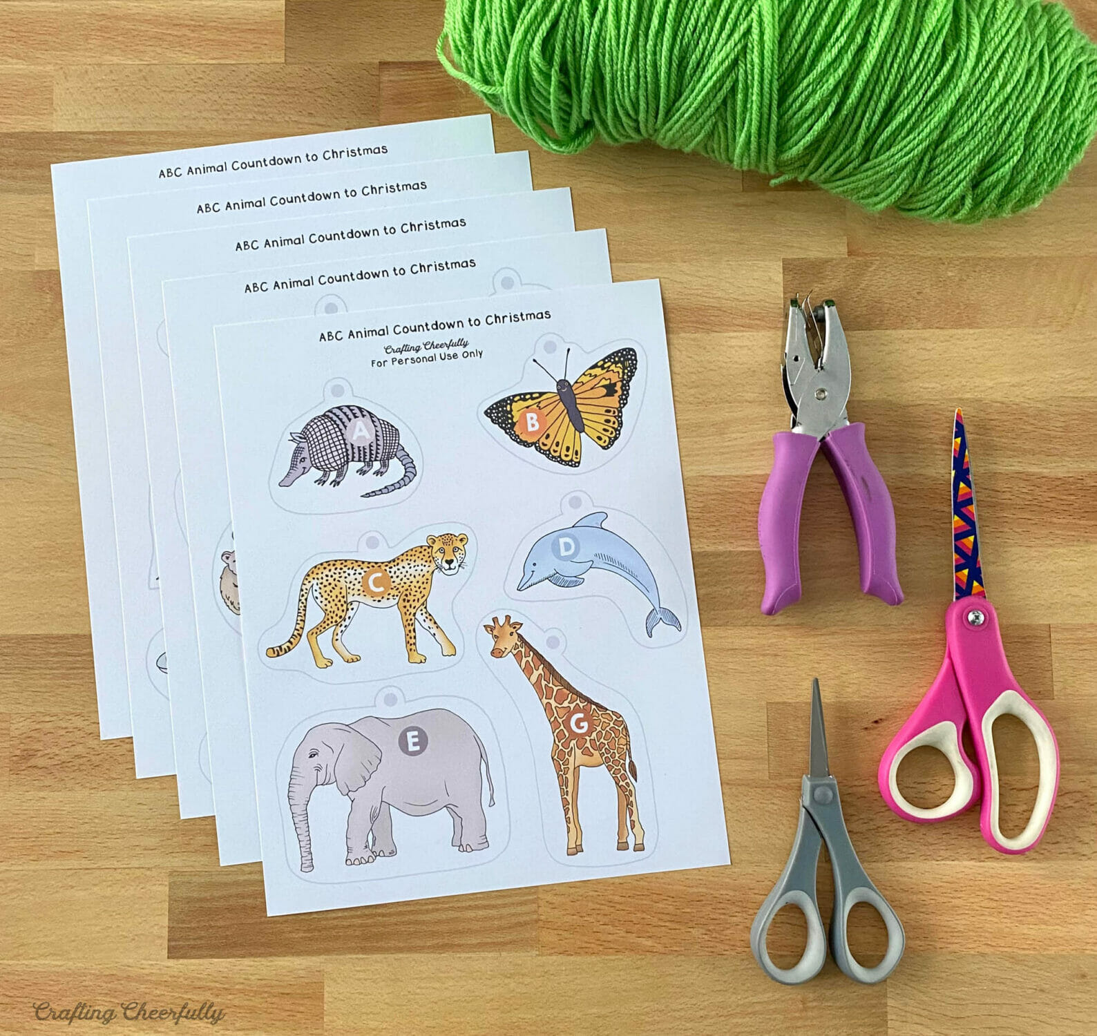 Pages of printable ornaments lay on a table with a scissors, hole punch and ball of green yarn.