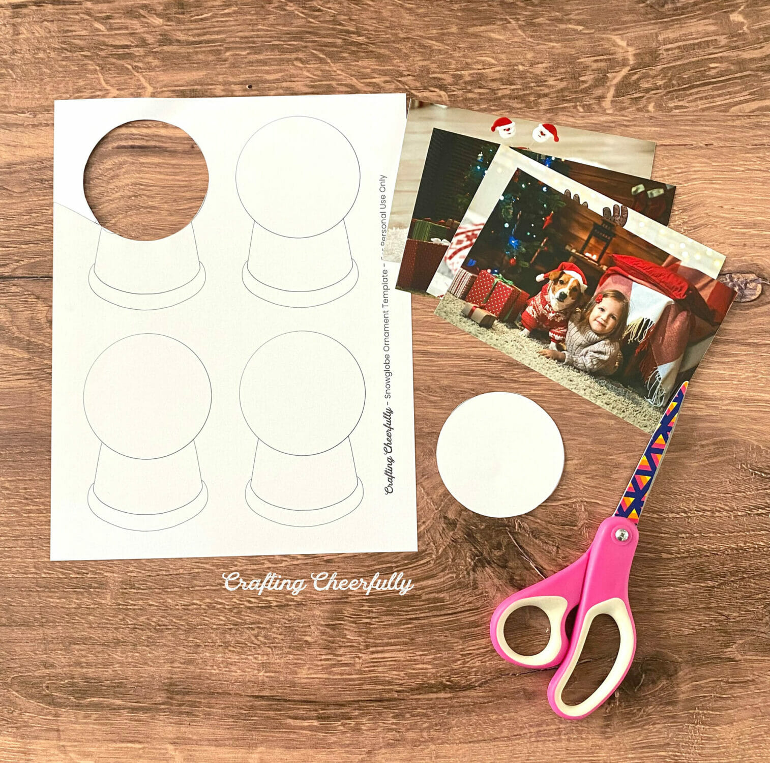 Snow globe template lays on a table next to a stack of holiday photos and a scissors.