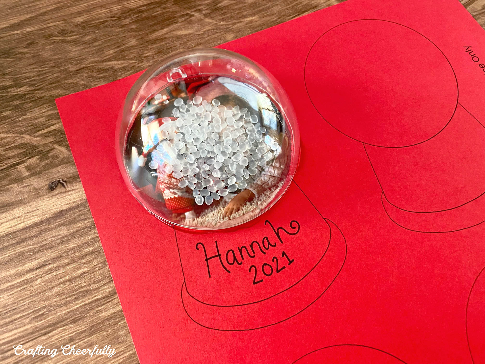 The template printed on red paper lays on a wooden table, It has a photo glued to it, poly-pellets lay on it and half of a plastic ornament is glued over top.