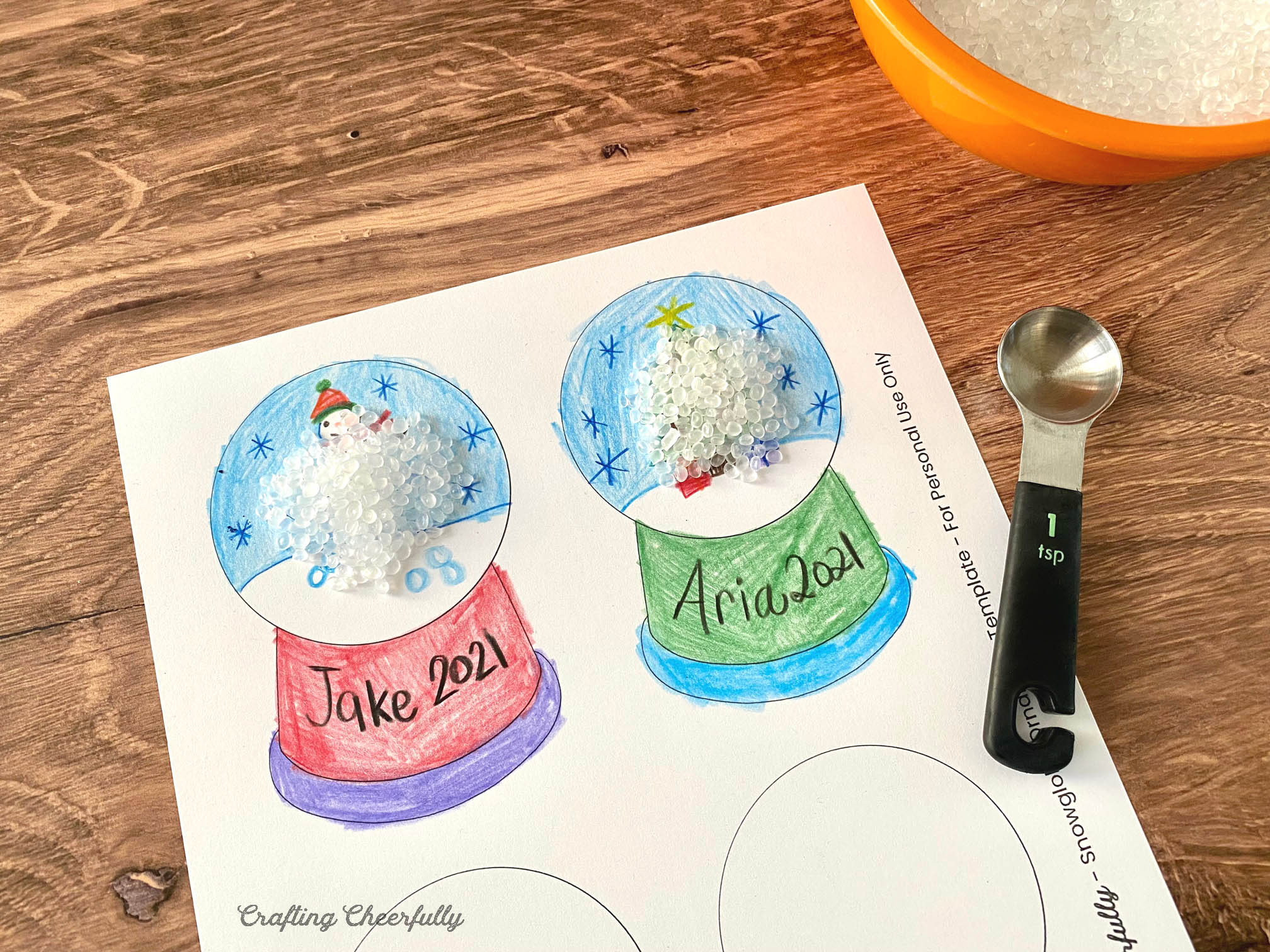 The snow globe template colored in lays on a wooden table with a bowl of poly-pellets and a teaspoon. 