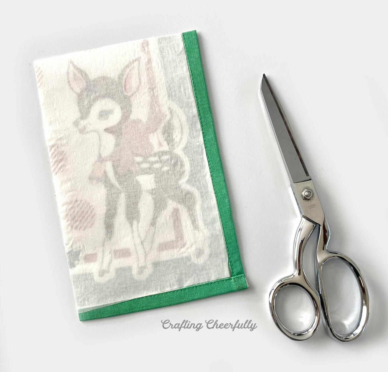 Fabric napkin with a deer printed on it laying next to a fabric scissors on a white table.