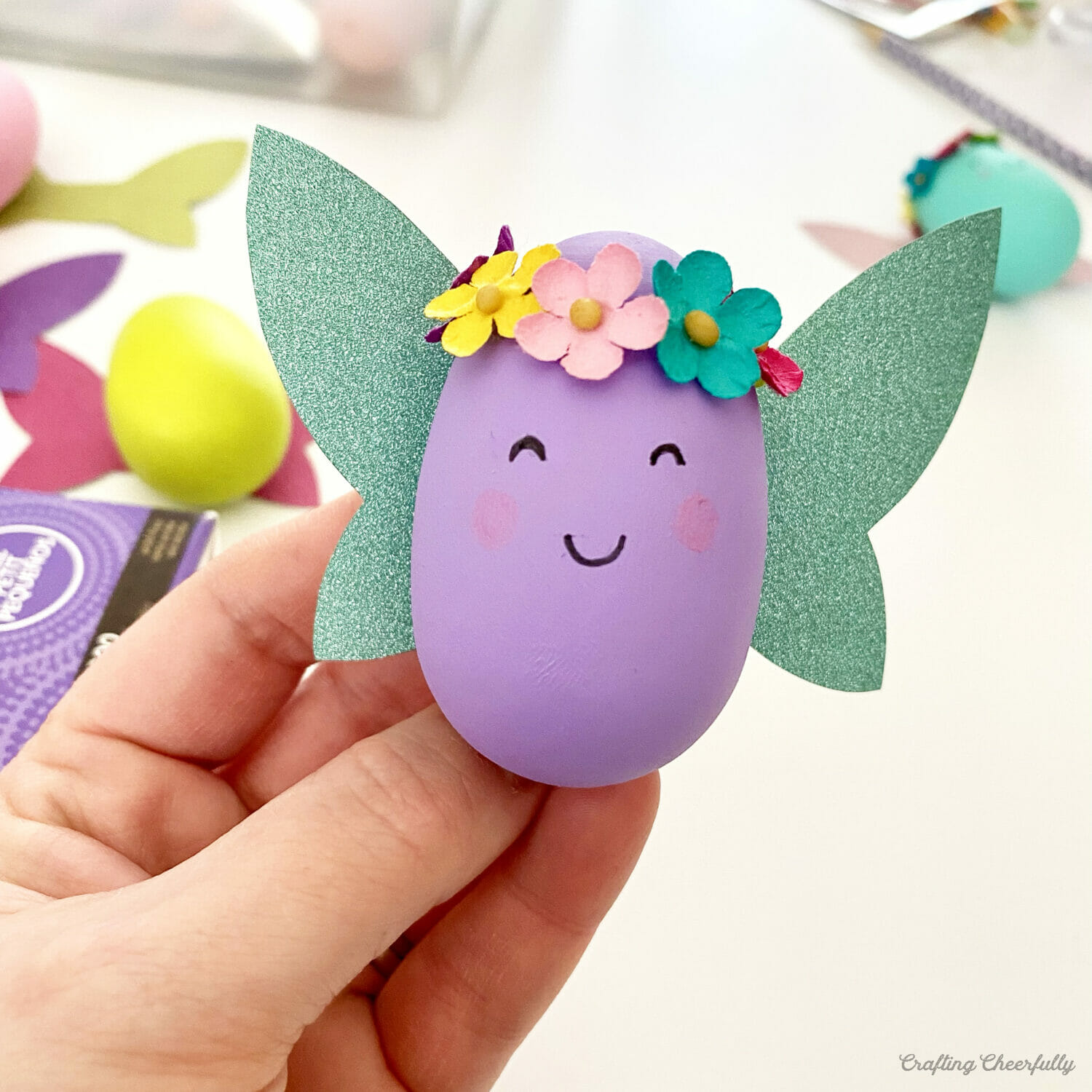 A hand holds up a sweet purple egg with a smiley face, glittery wings and flower headband.