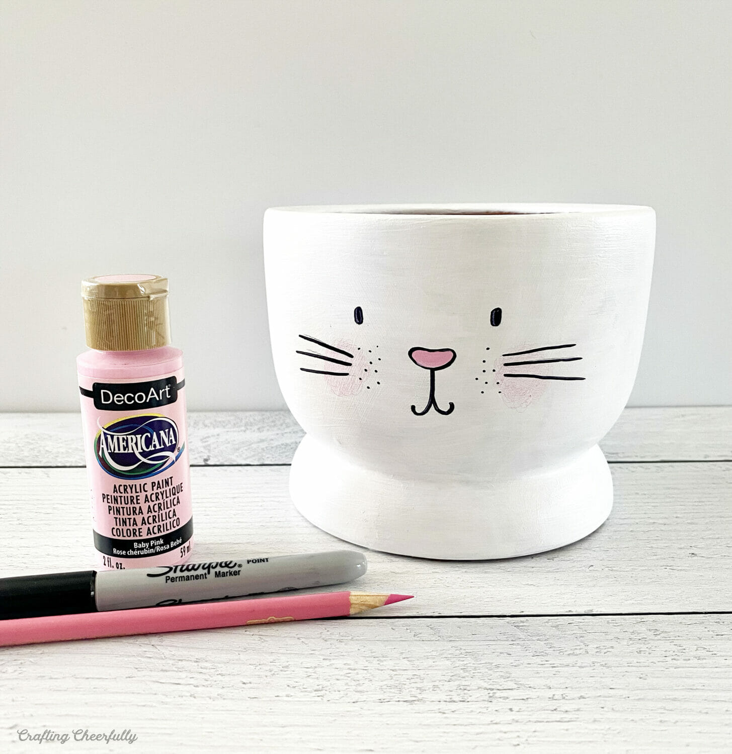 A cute bunny face on a white pot with a bottle of pink paint, pink colored pencil and black Sharpie marker.