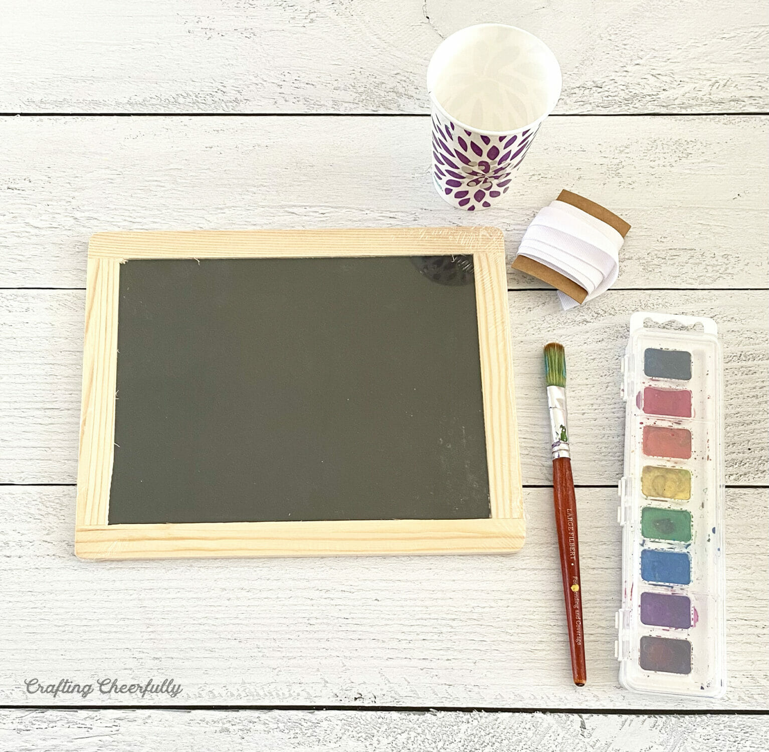 Supplies needed for creating the chalkboard sign including a mini chalkboard, water cup, watercolor paint, paint brush and ribbon.