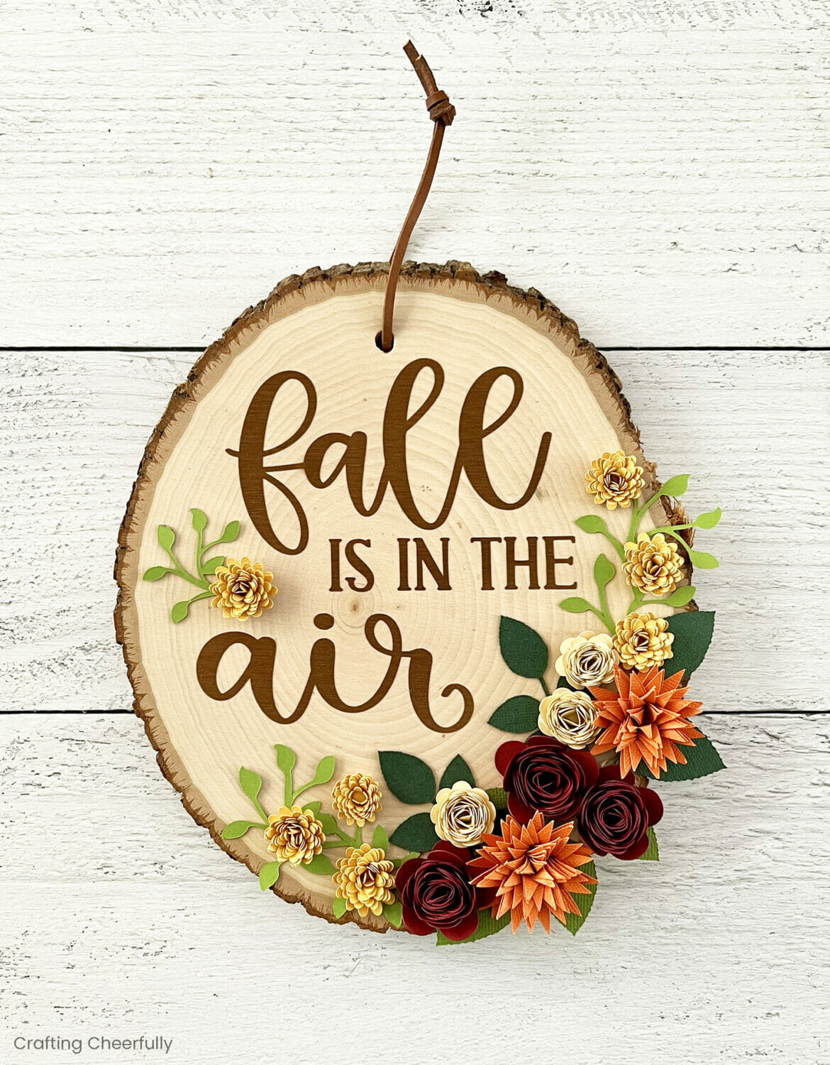 Wood slice decorated with paper flowers in autumn colors and the phrase fall is in the air.