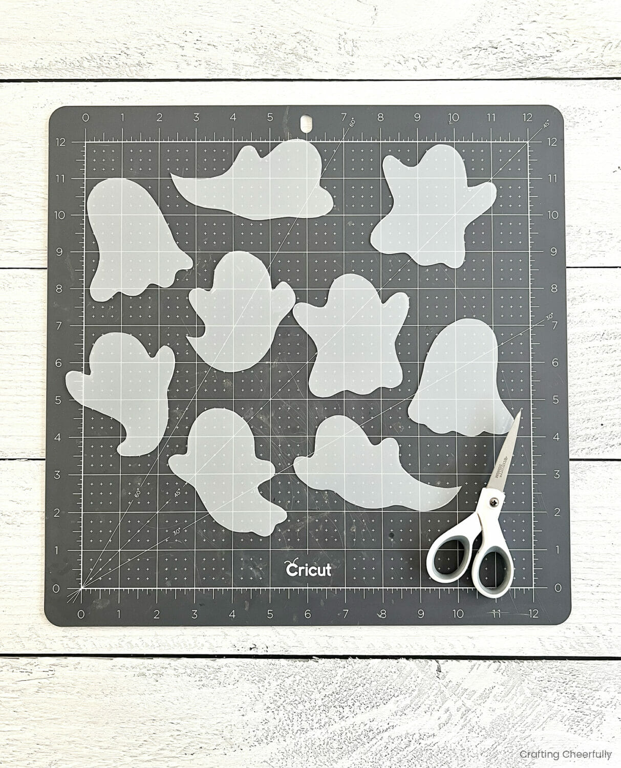 Lots of ghost shapes cut from craft plastic lay on a black cutting mat next to a scissors.