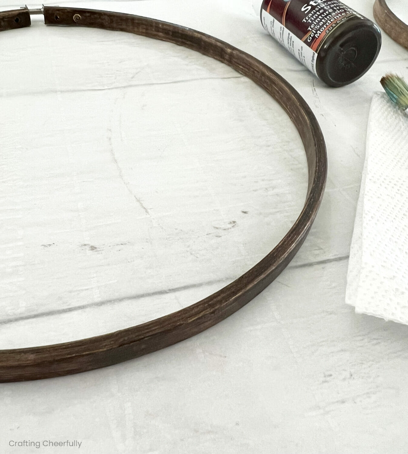A hoop that is being stained lays next to stain, paper towel and a paint brush.