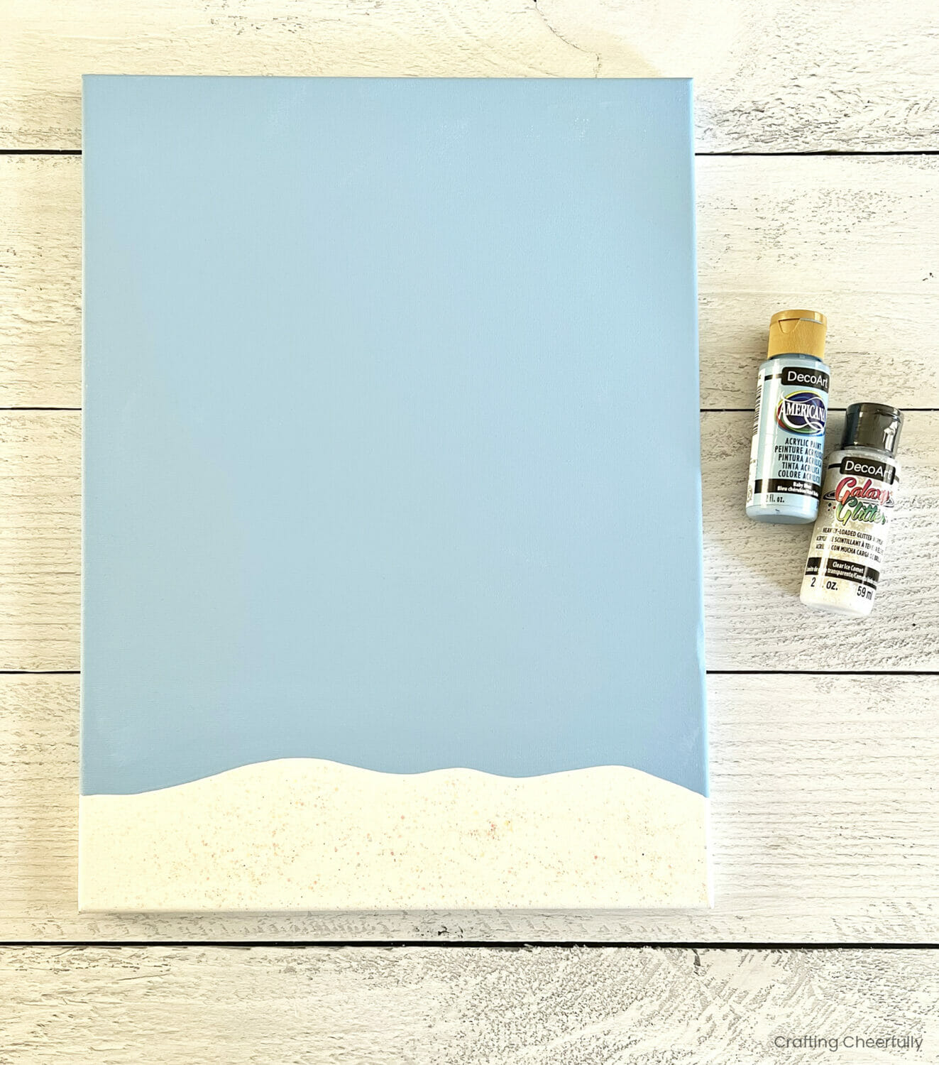 A canvas is painted to have a blue sky and snow at the bottom using acrylic paint. 