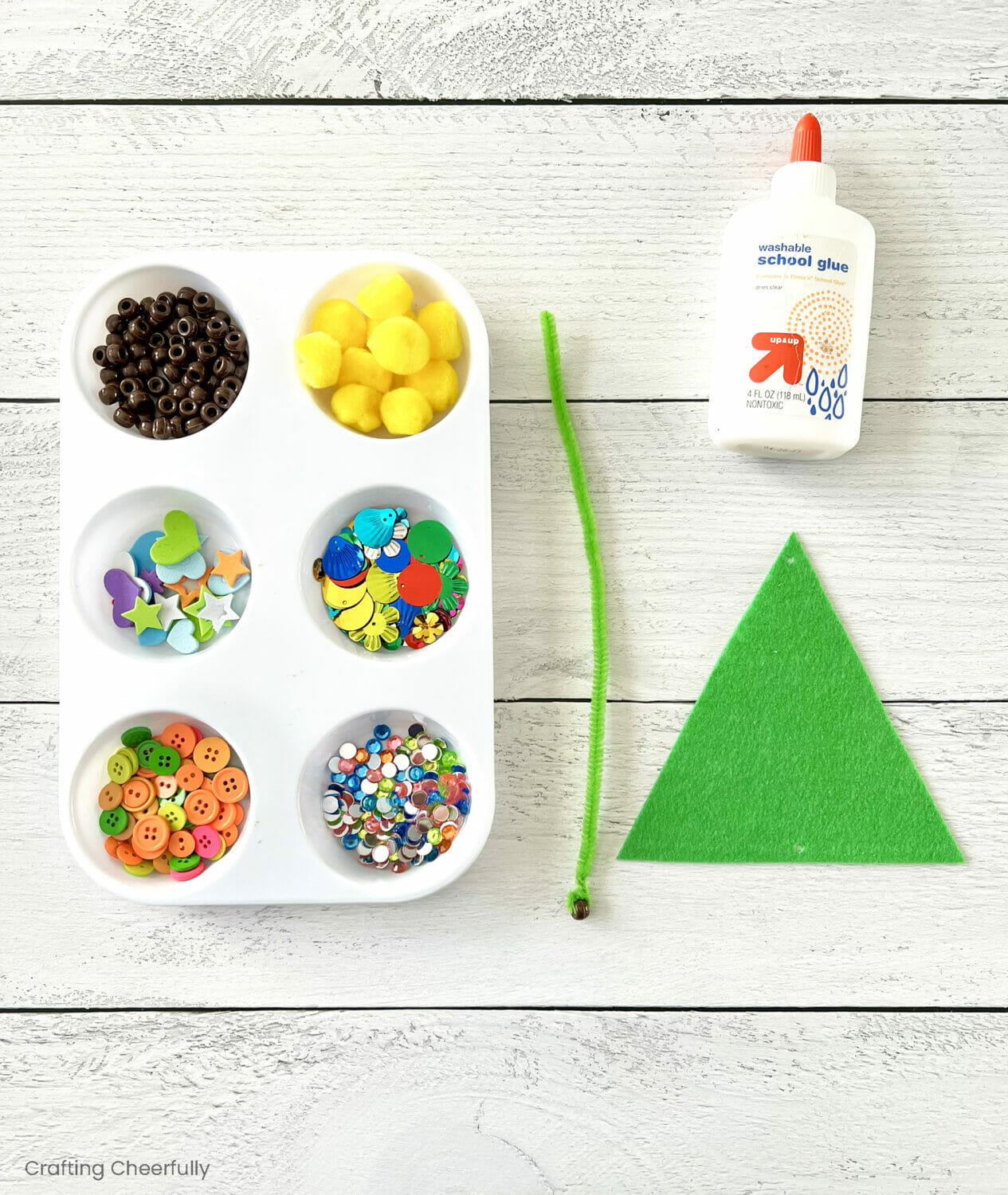 A tray of decorating supplies lays next to a bottle of glue, green triangle and pipe cleaner.