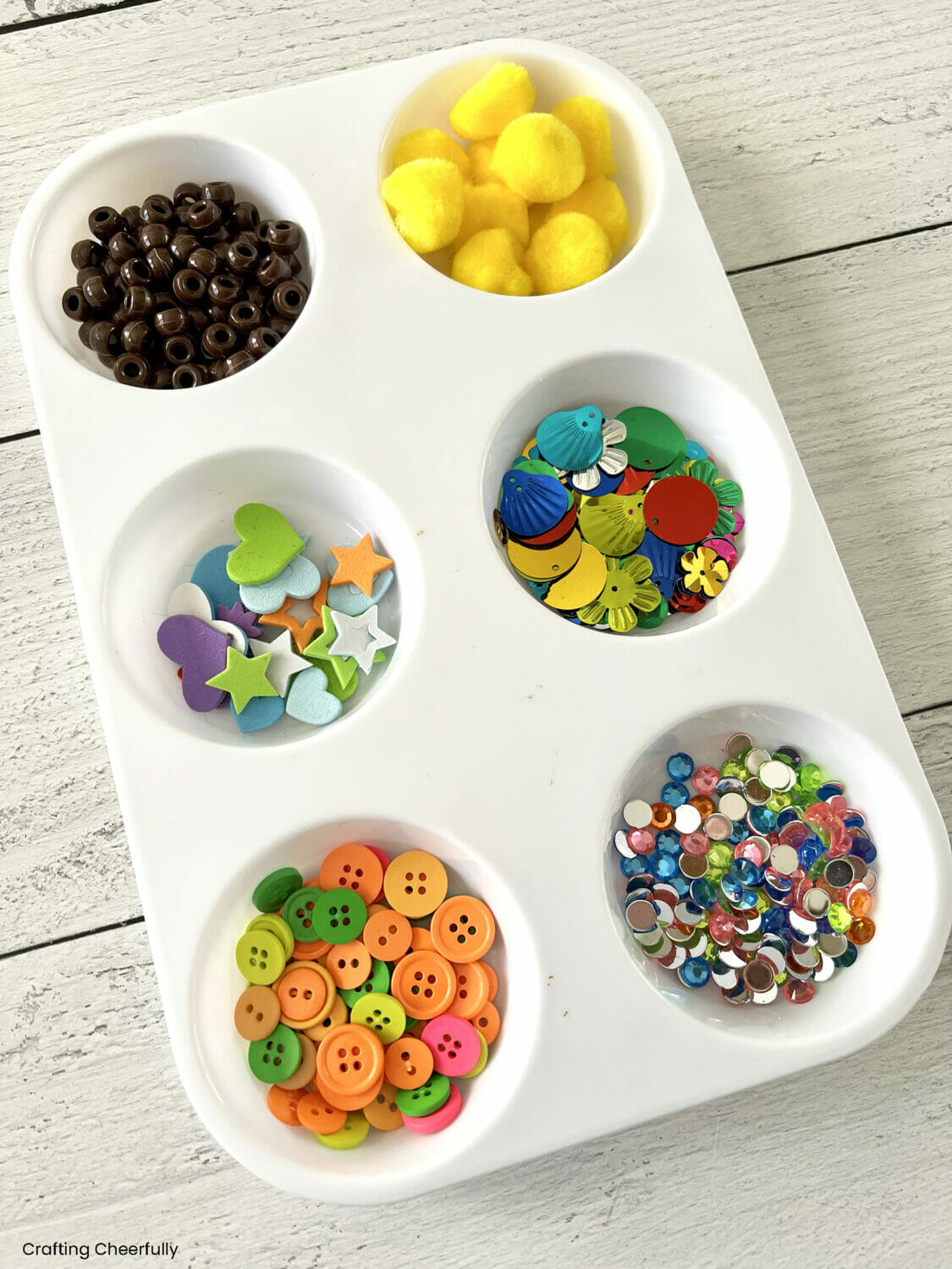A plastic tray filled with buttons, foam stickers, sequins, gems, yellow pom poms and brown pony beads.