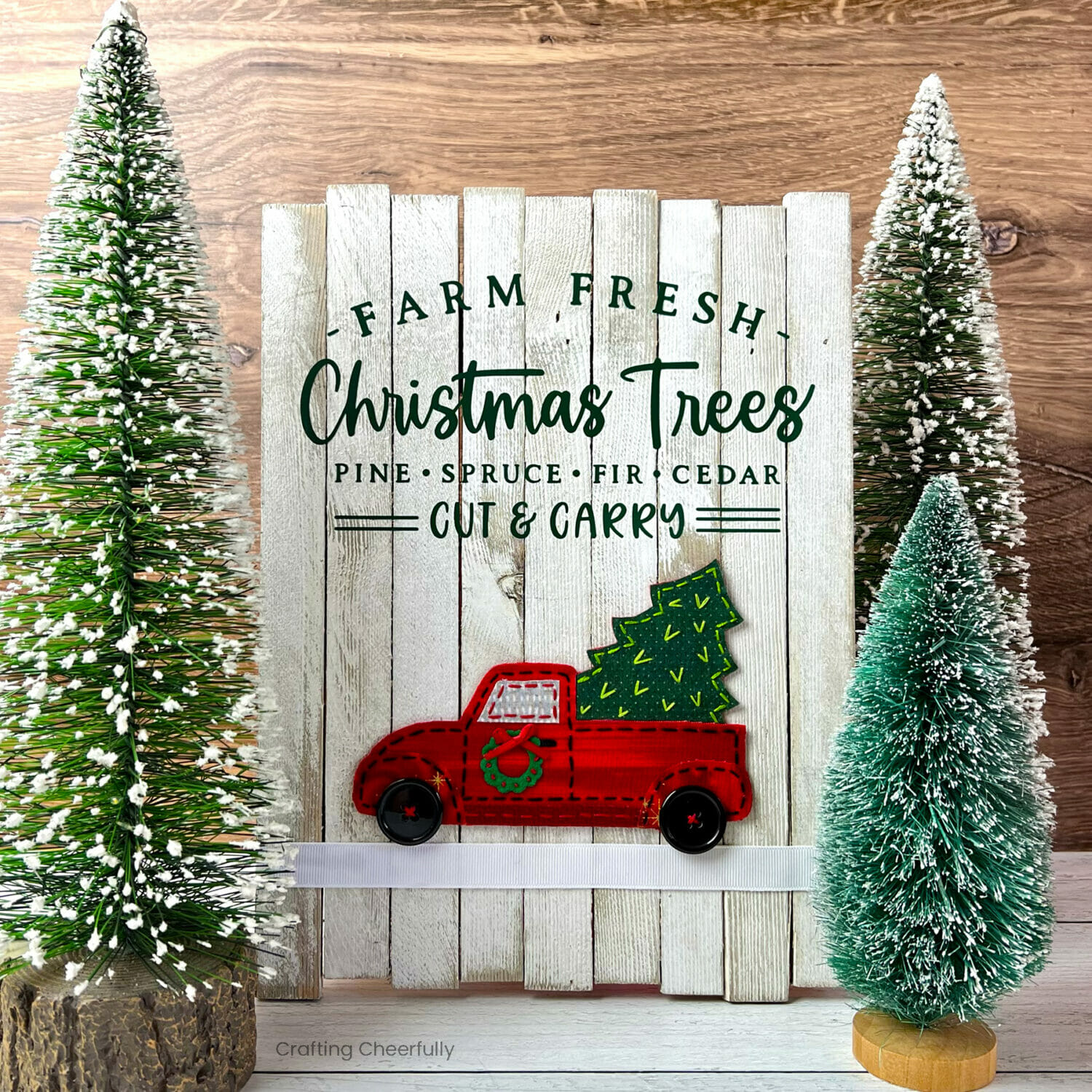 White wooden Christmas sign that reads "Farm Fresh Christmas Trees" with a fabric red truck on it sits next to bottle brush trees.