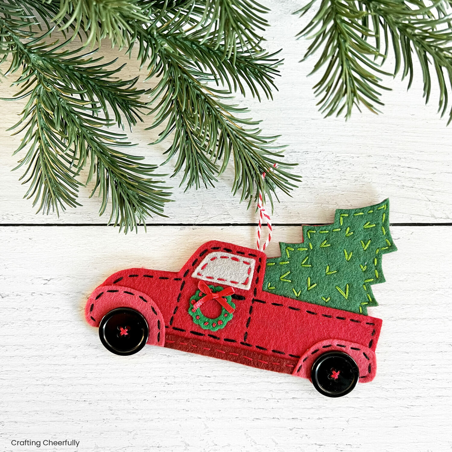 A vintage red truck ornament made out of felt hangs from an evergreen branch.