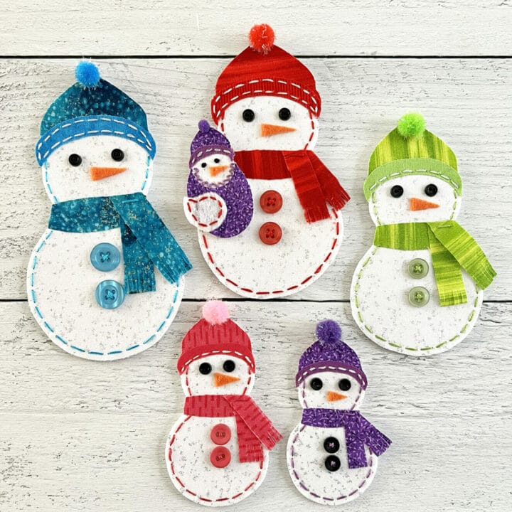 A group of fabric snowmen lay on a wooden table with bright and colorful hat and scarves.