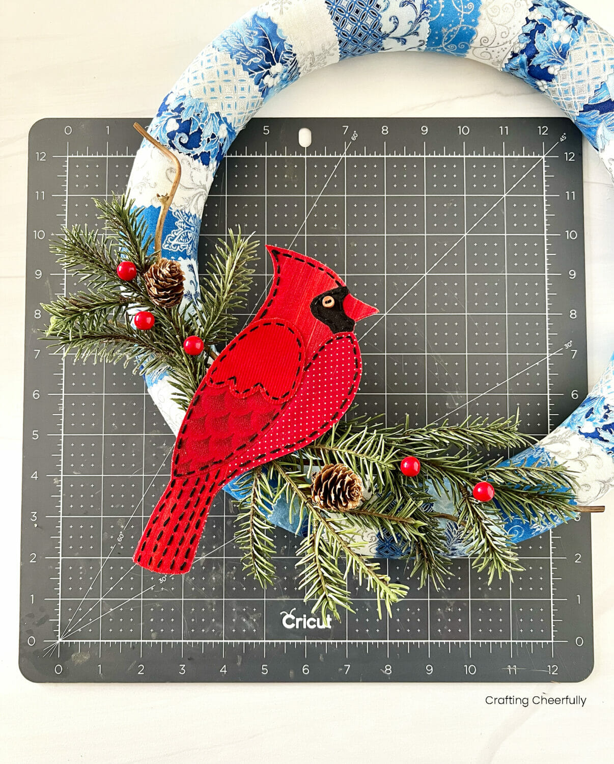 A red fabric cardinal sits on top of pine branches on the bottom of a wreath.