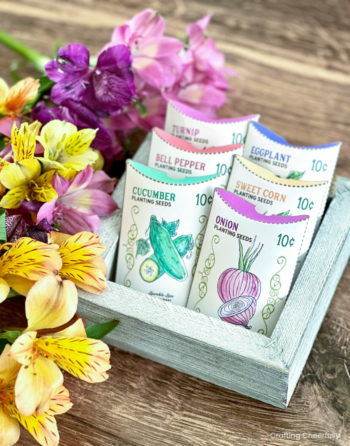 Vegetable seed treat boxes sit in a wooden box next to flowers.