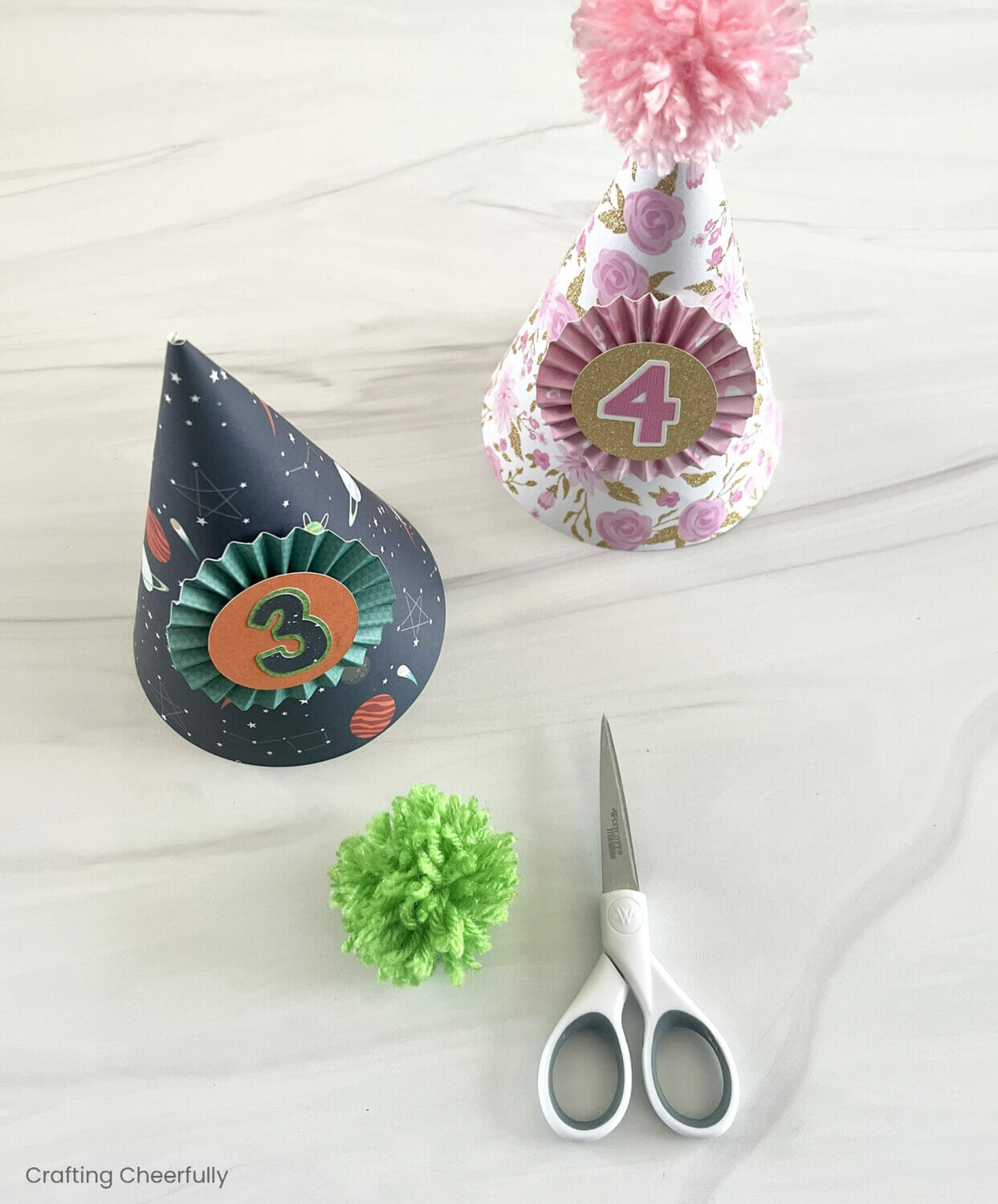 Two paper party hats; one with a pink pom pom and the the other about to get a green pom pom.