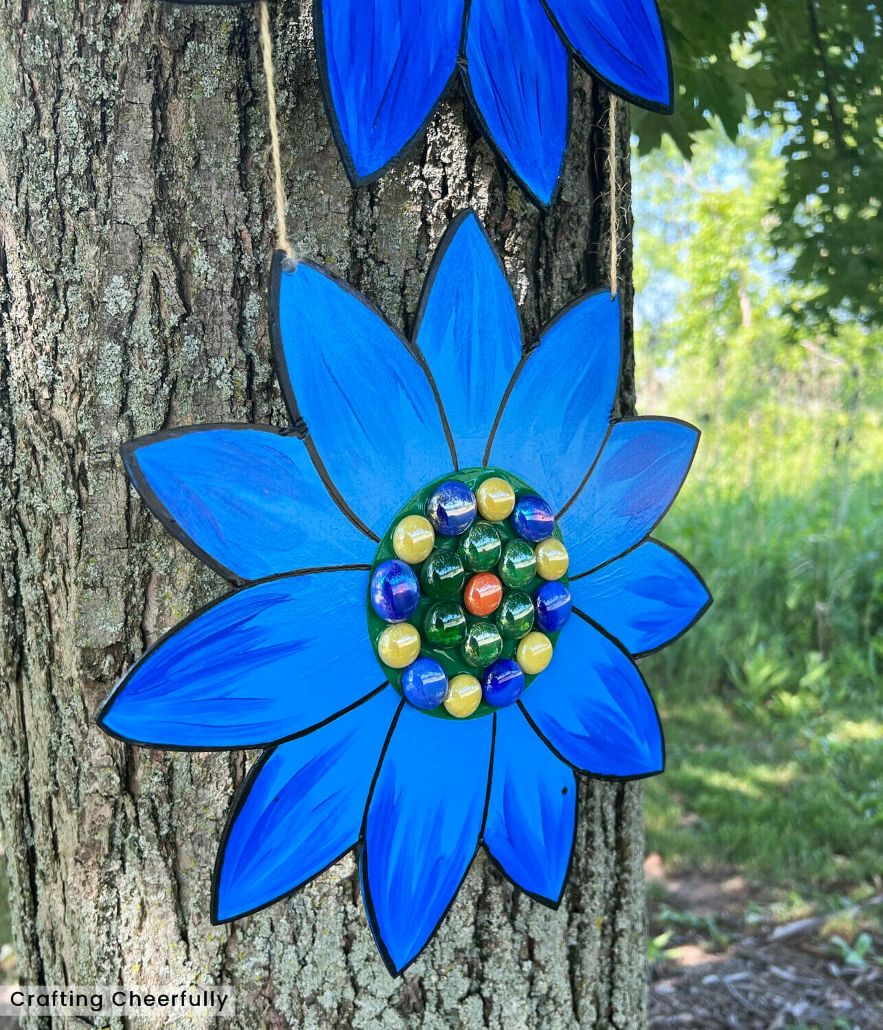 Blue wooden flower hanging in a tree with colorful gemstones at the center of the flower.