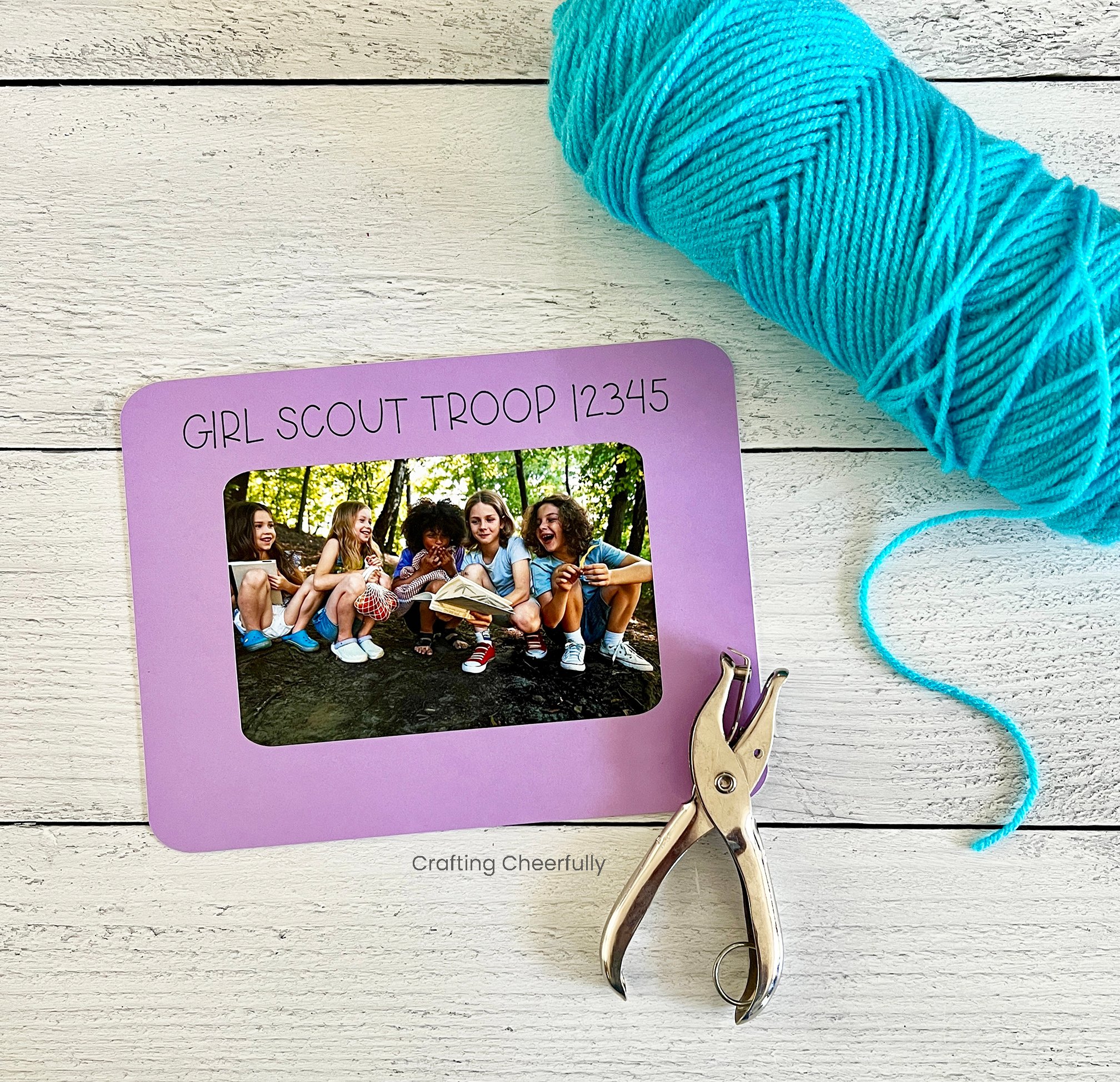 A purple photo frame that reads Girl Scout Troop lays next to a hole punch and a roll of yarn.