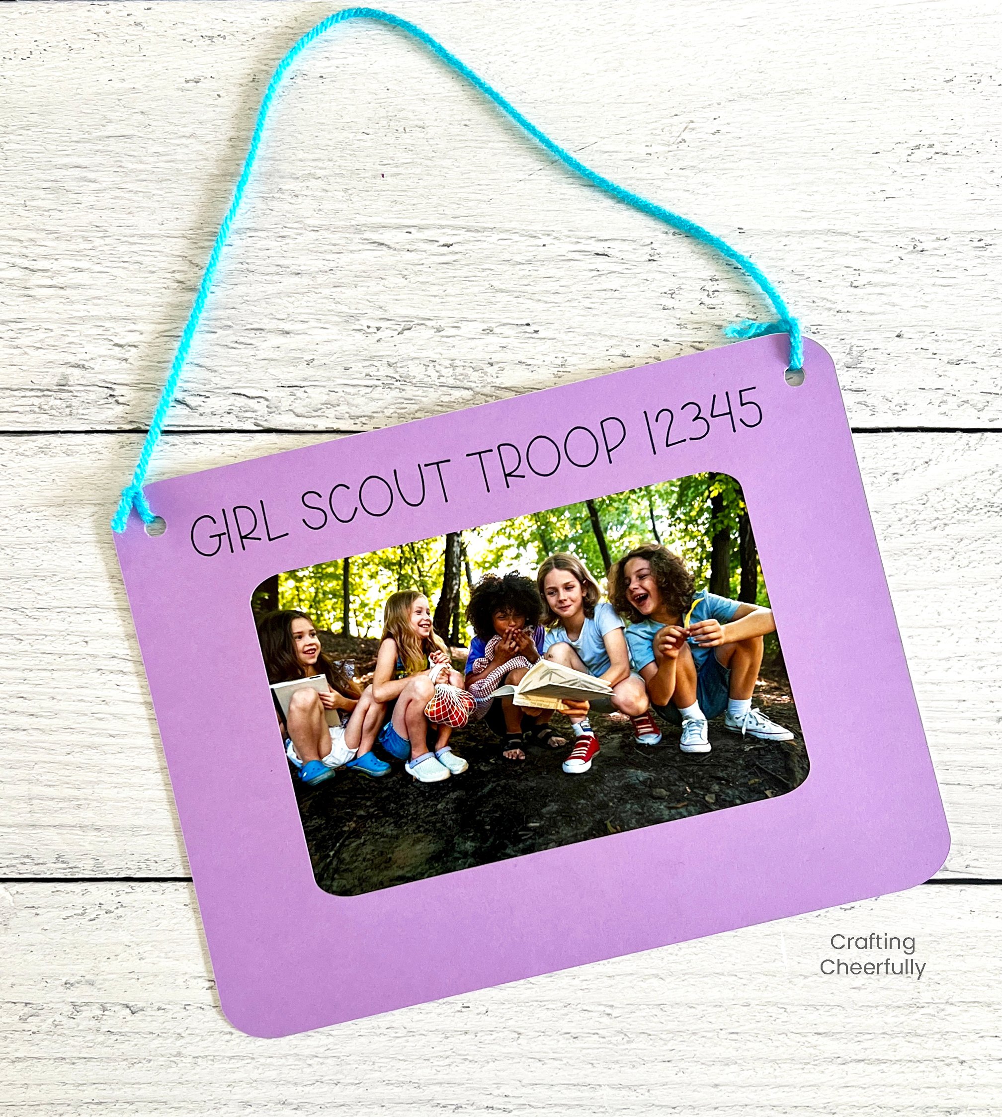 A purple photo frame that reads Girl Scout Troop has a blue yarn hanging thread.