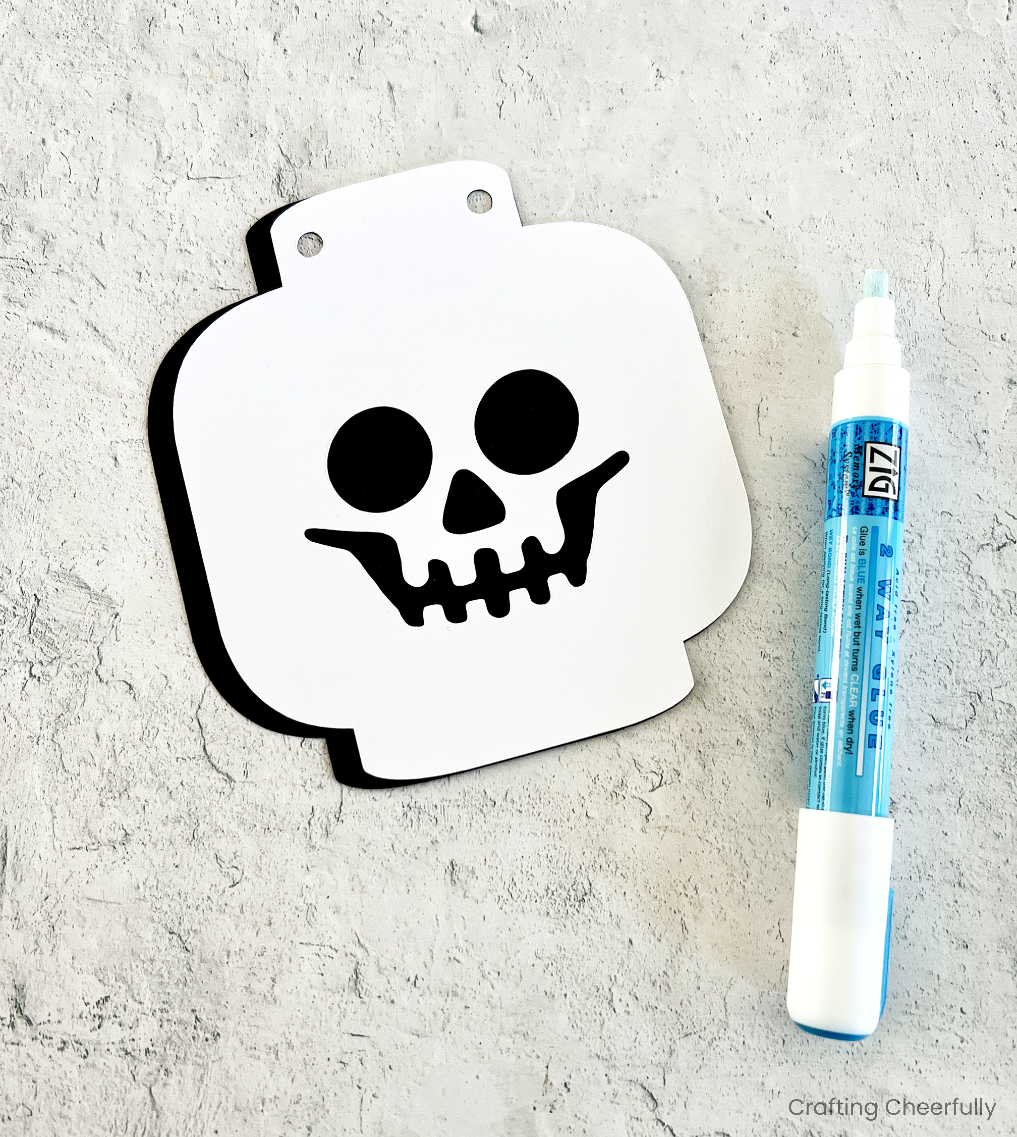 A white lego skeleton face with cutouts for the eyes, nose and mouth lays on top of a black piece of paper next to a glue pen.