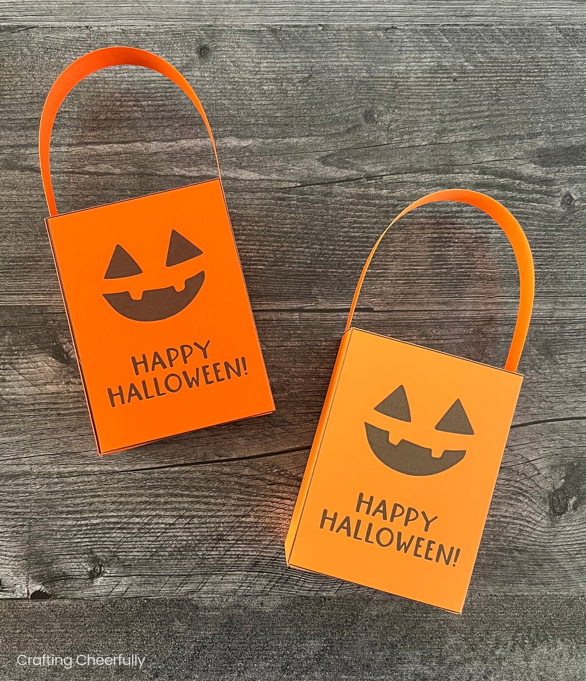 Two pumpkin treat boxes with handles read Happy Halloween lay on a black wooden surface.