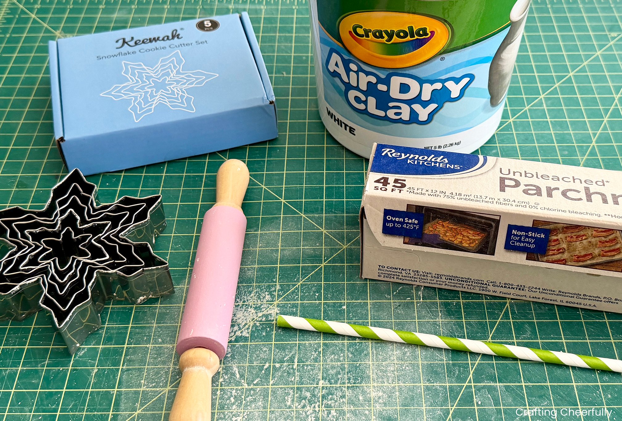 Supplies including rolling pin, snowflake cookie cutters, parchment paper, air-dry clay, and a straw on a self-healing cutting mat.