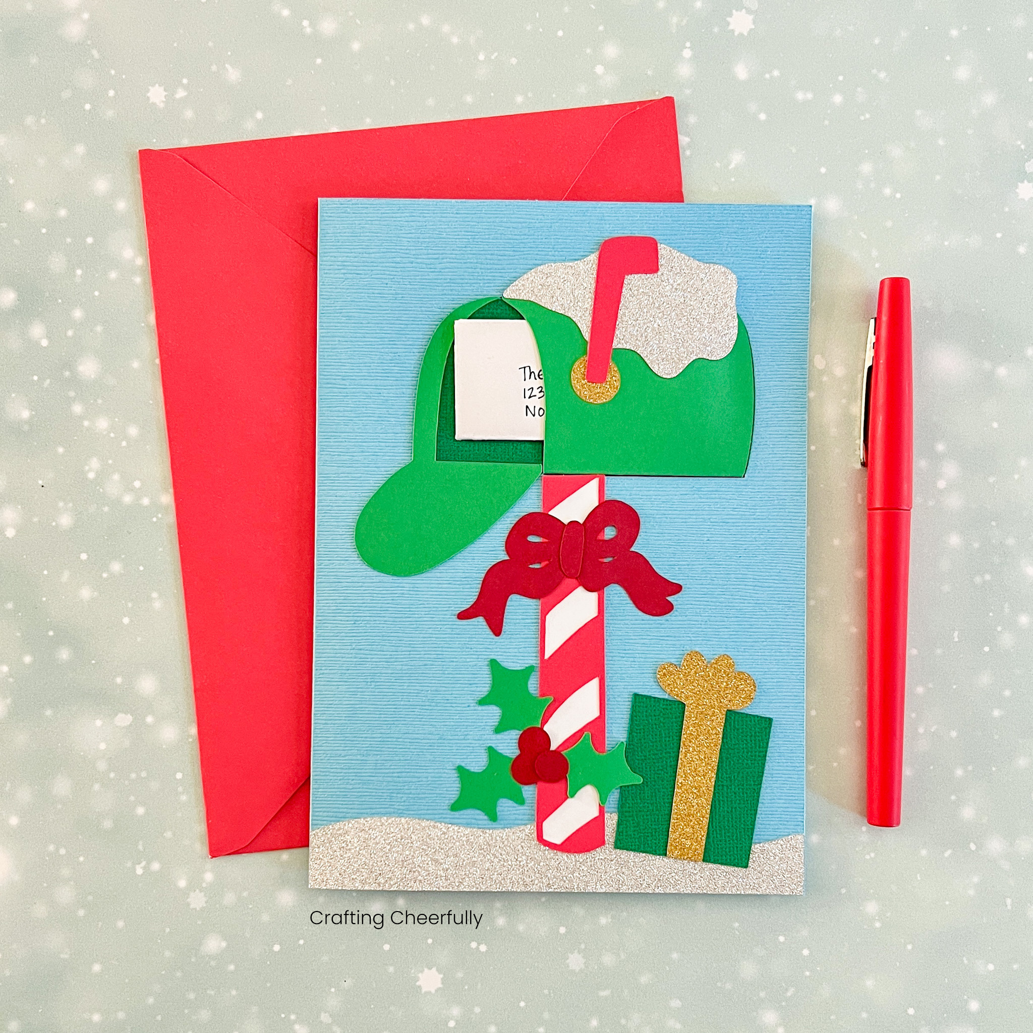 A Christmas holiday mailbox card has a festive mailbox on the cover created from paper; an envelope is seen peeking from inside the mailbox.