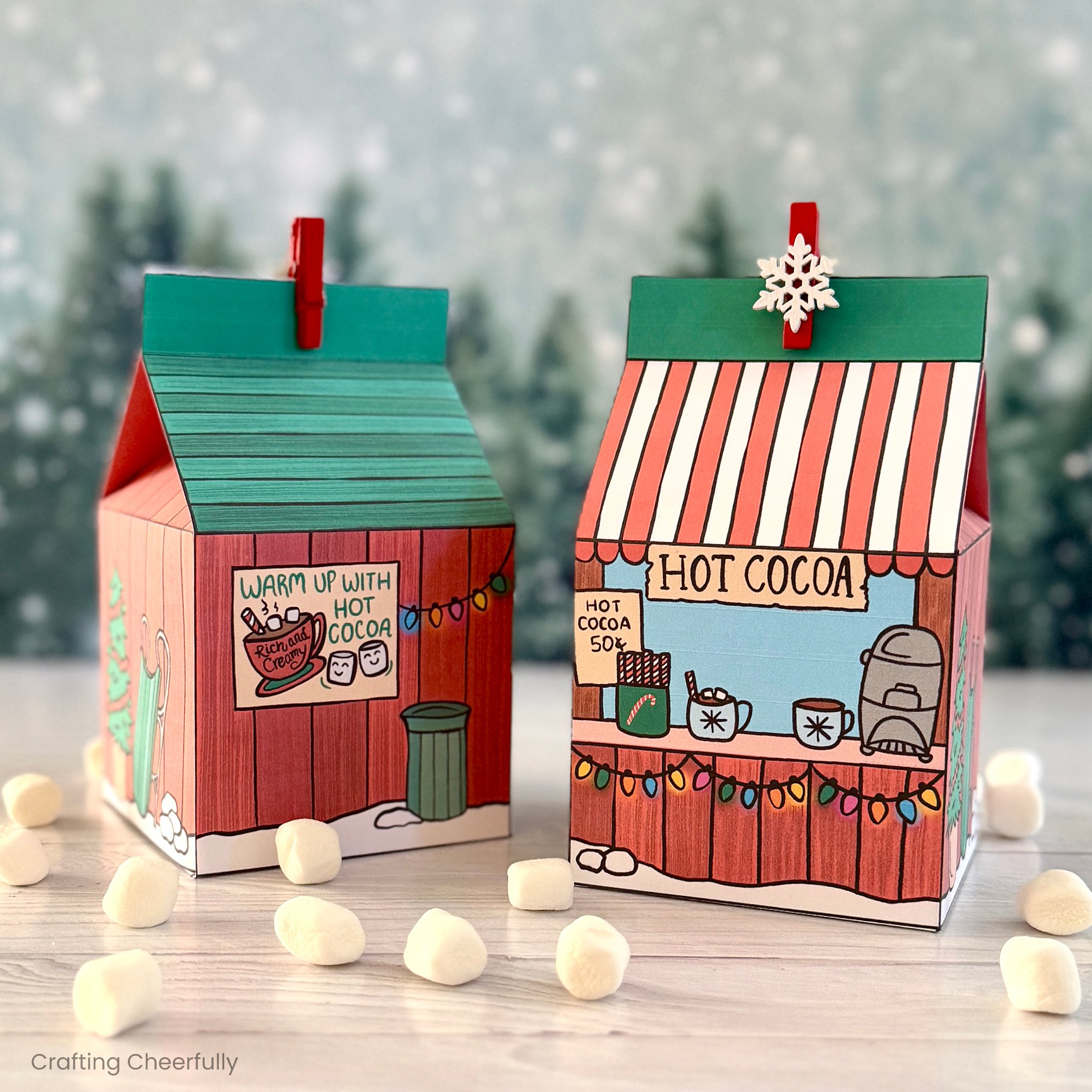 Printable hot cocoa treat boxes sit on a wood table with tiny marshmallows around them.
