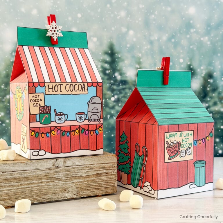 Printable hot cocoa treat boxes sit on a wood table with tiny marshmallows around them.