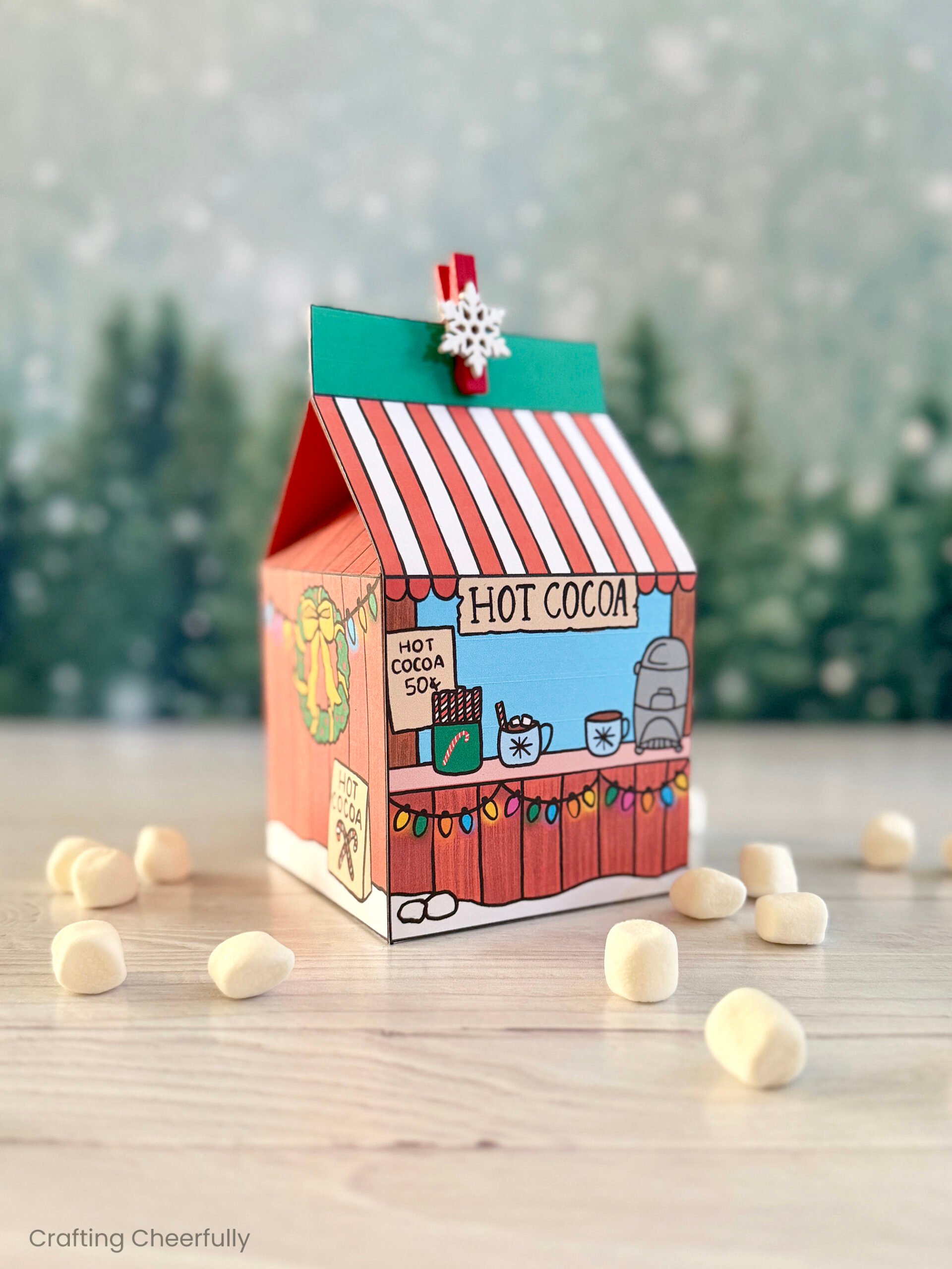 A printable hot cocoa treat box sits on a wood table with tiny marshmallows around them with evergreen trees in the background.