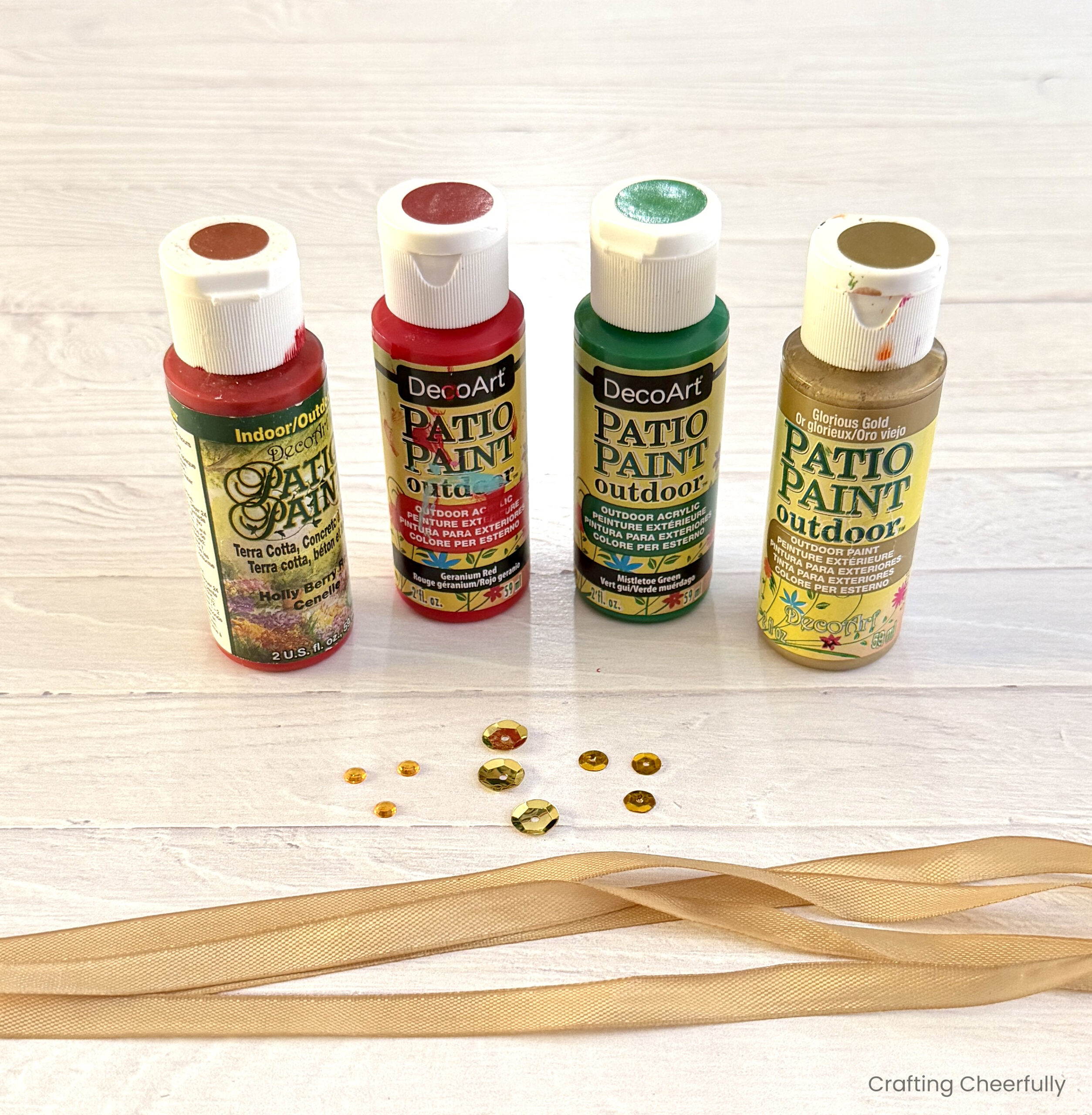 Four bottles of paint, gold sequins and gold ribbon lay on a white table.