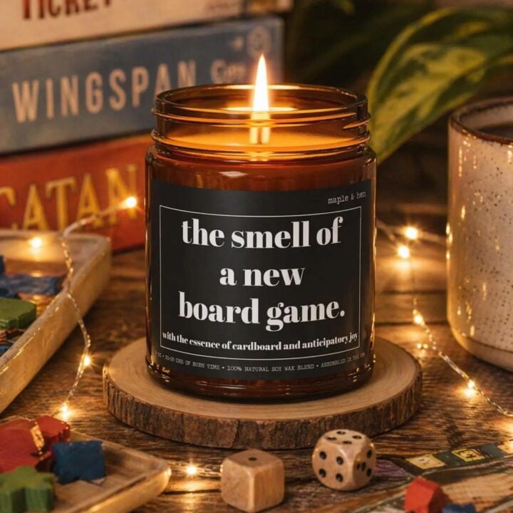 Candle called "The smell of a new board game" sits with board games and dice. Created by maple and Hen Etsy Shop.