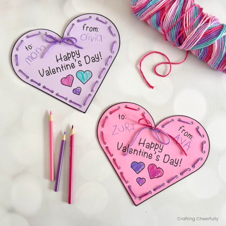 Two heart valentines that have been laced with pink and purple yarn lay on a table with a ball of yarn and colored pencils.