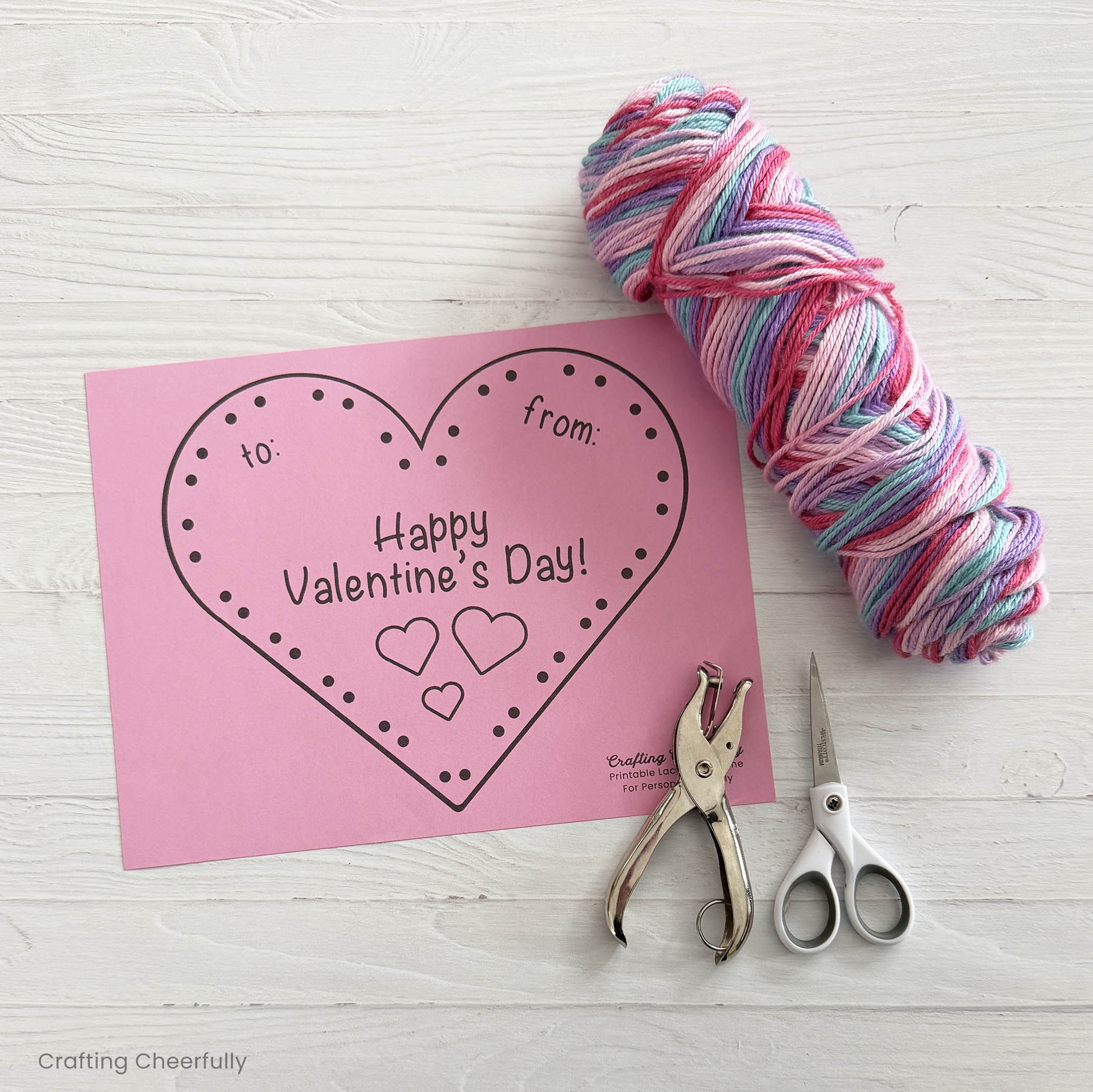The supplies needed for the valentine craft are gathered on a table including the printable, hole punch, scissors and yarn.