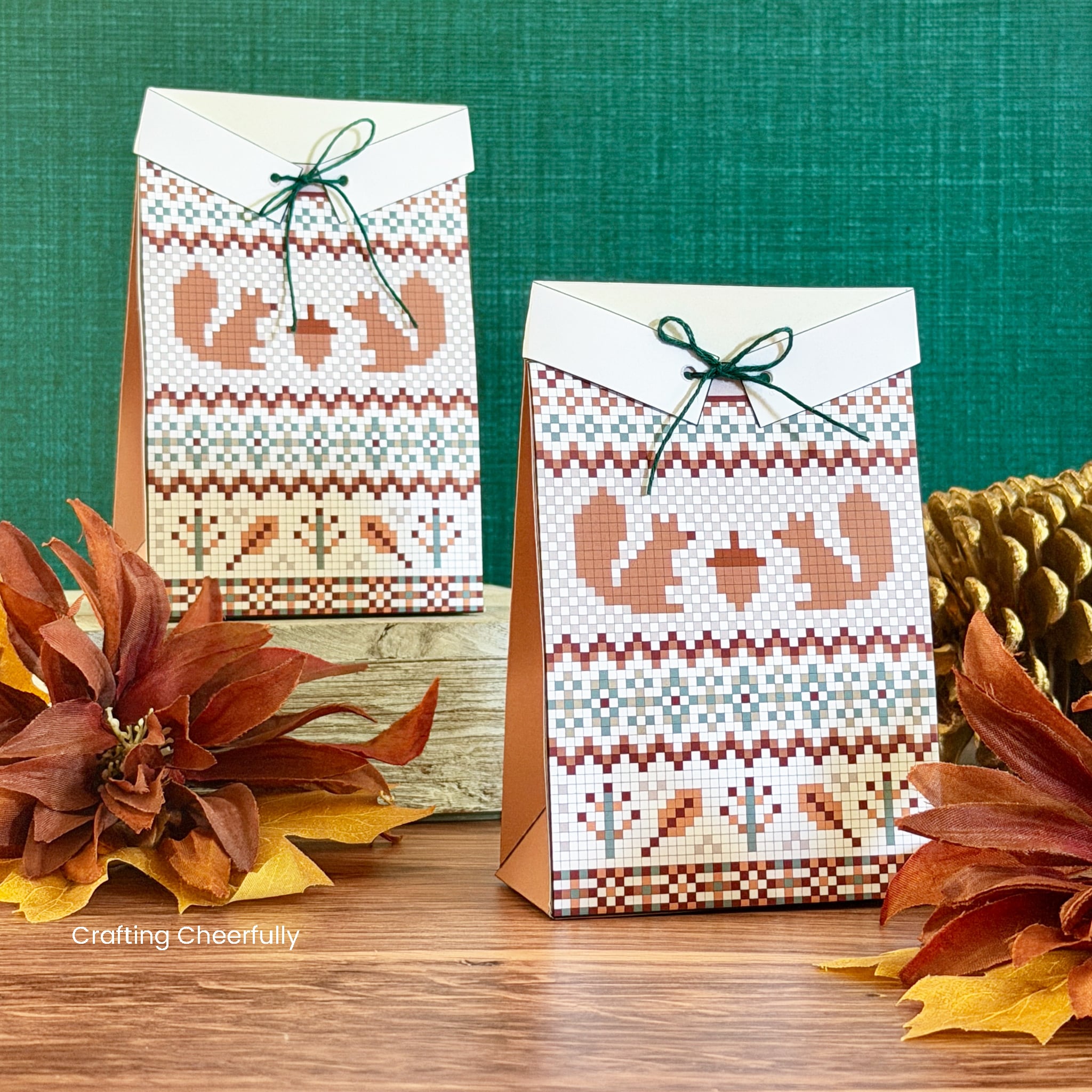 Two paper treat boxes designed to look like fall swaters with a squirrel and acorn design on the front sit on a table with pinecones and fall leaves next to them.