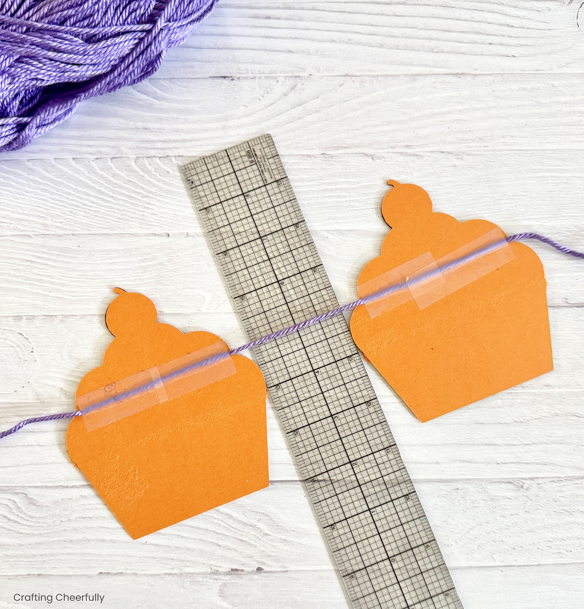 A ruler lays between two paper cupcakes as a string of yarn is taped to the back of each cupcake.
