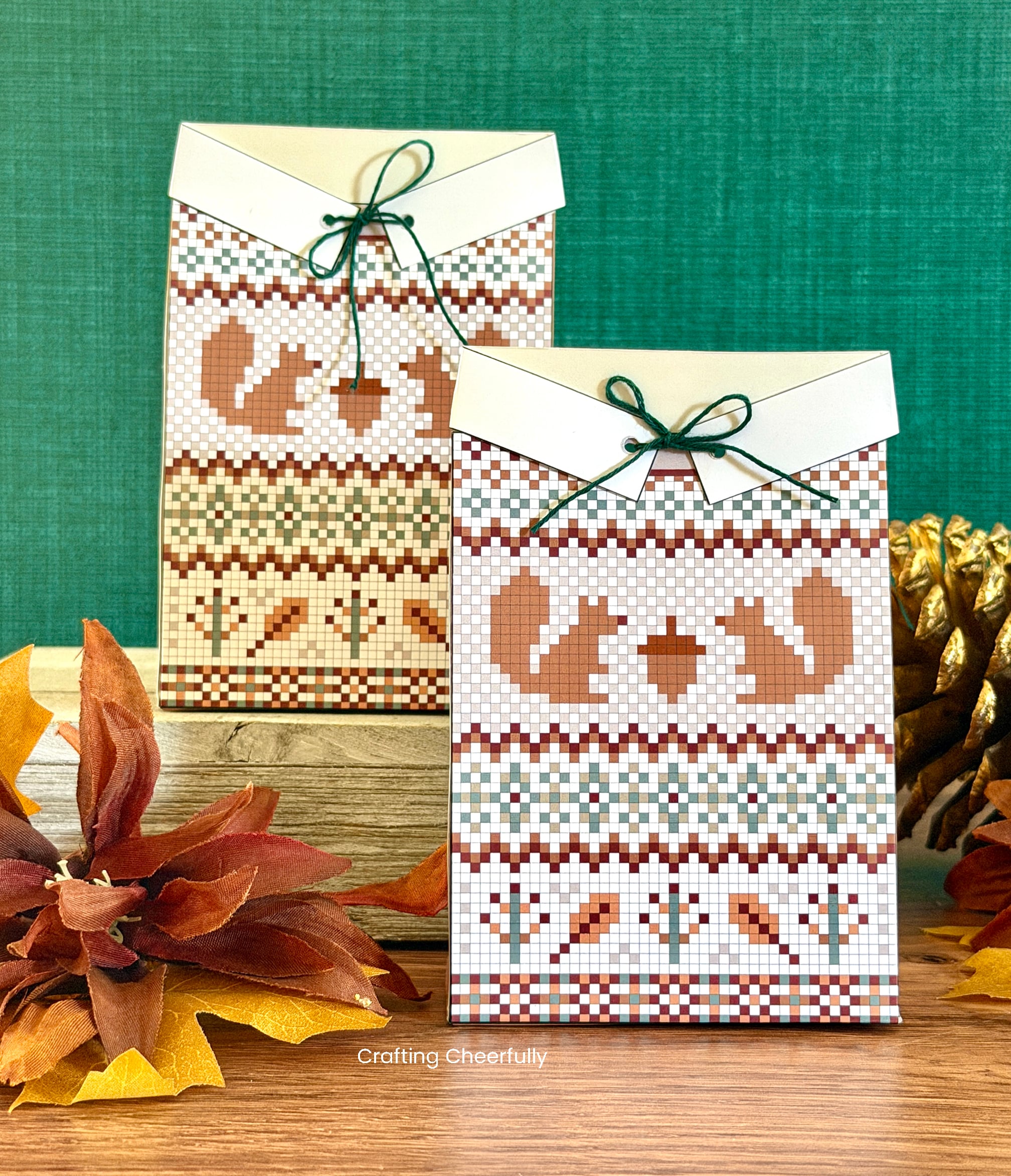 Two paper treat boxes designed to look like fall swaters with a squirrel and acorn design on the front sit on a table with pinecones and fall leaves next to them.